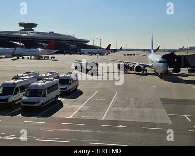 Large blue airplane is parked on the tarmac at an airport. There are several other airplanes in the background, and a number of vehicles, including a bus and a truck, are parked around the airport Stock Photo