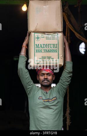 A worker in a green long sleeve shirt and a red head cloth balances boxes on his head inside a dark warehouse lit by a single bulb, West Bengal, Howra Stock Photo