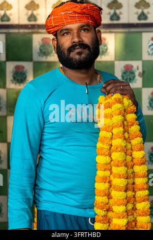 Close portrait of a flower seller in a turquoise long sleeve shirt posing with a heavy strand of yellow and orange blossoms against floral tiles at th Stock Photo