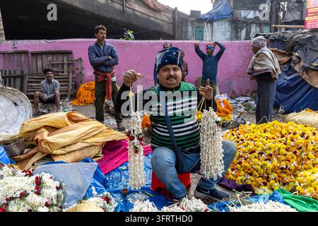 Man in a striped sweater and blue woolly hat sits on a stool showing long white flower strands near yellow marigold heaps and a pink wall at the flowe Stock Photo