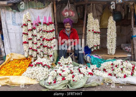 A man in a red jacket and purple headcloth sits in a small stall framed by hanging white and red garlands with marigolds and jasmine at the flower mar Stock Photo