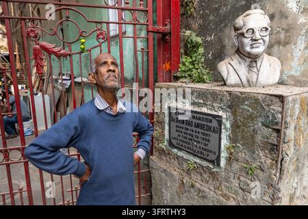 Elderly man in a collared shirt stands next to a monument with a white bust of a bespectacled man on a weathered pedestal by an ornate red iron gate, Stock Photo