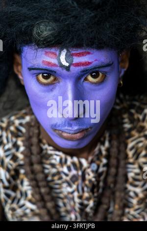 Close portrait of a man disguised as the Hindu god Shiva with violet blue face paint red stripes a white crescent and intense yellow eyes, West Bengal Stock Photo