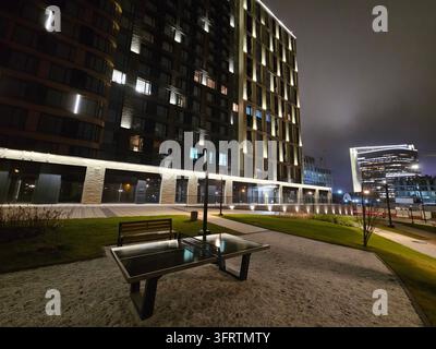 Nighttime view of modern building with illuminated windows. Outdoor bench sits on gravel path beside green lawn. Building facade combines stone and glass with warm lighting Stock Photo