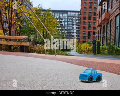 Blue toy truck sits on gravel path. Modern buildings loom in background. Autumn leaves frame the scene. Play structure stands nearby. Urban park setting with trees. Bright day enhances colors Stock Photo