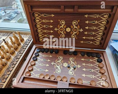 Wooden backgammon set with intricate inlay designs. Black and tan checkers arranged on patterned board. Game pieces and dice placed on playing surface Stock Photo