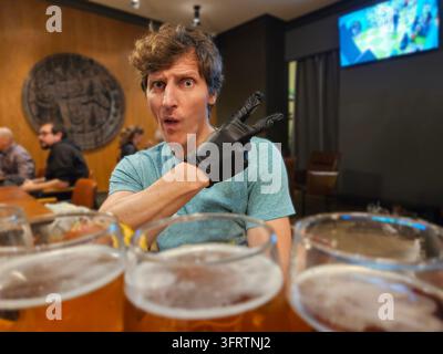 Man gestures with gloved hand at beer glasses. He sits at a table in a pub setting. Background shows patrons and a large round emblem. Beer glasses fi Stock Photo
