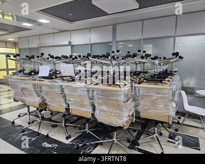 Modern office chairs lined up in rows. Plastic wrap covers most of the chairs. Metal frames and wheels are visible. Chairs sit on a tiled floor surface. Background shows glass-walled office space Stock Photo