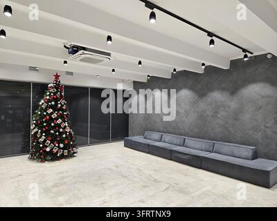 Modern minimalist room with Christmas tree and sofa. Dark concrete walls contrast with white ceiling and floor. Track lighting highlights the space sleek, clean lines Stock Photo