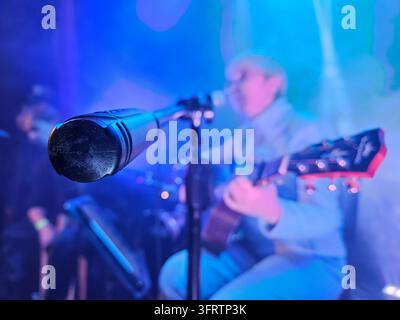Blue-lit stage with microphone in foreground. Musician plays guitar softly in background. Stage haze and spotlights create atmospheric glow. Focus on mic, blurred performer adds depth Stock Photo