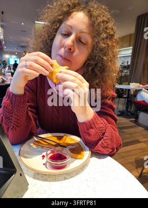 Woman enjoys snack at restaurant table. Curly hair, red sweater, hands holding food. Plate holds fries and red sauce. Background shows casual dining area. Natural lighting, relaxed atmosphere Stock Photo