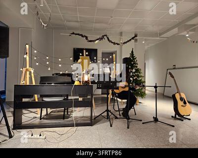 Musician plays piano and guitar in festive room. Strings lights decorate ceiling and walls. Christmas tree stands near instrument stands. Guitar rests on stand beside keyboard Stock Photo