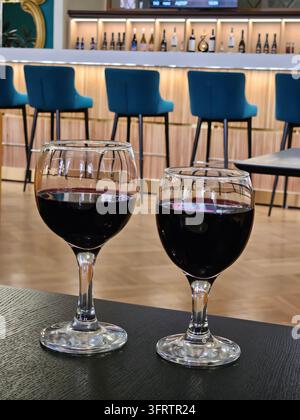 Two wine glasses filled with red wine sit on a dark table. Bar with blue stools is visible in the background. The setting is warm and inviting, with soft lighting. The glasses are clear Stock Photo