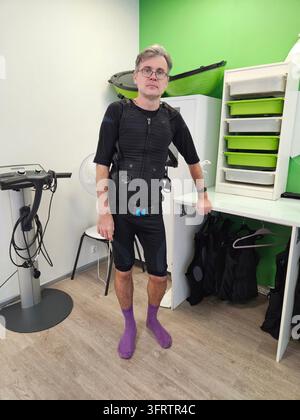 Man wears black resistance suit and purple socks. He stands in fitness studio with walls. Equipment and storage units are visible behind him. He looks directly with glasses electro muscle stimulation Stock Photo