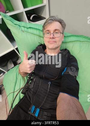 Man gives thumbs-up while wearing electric muscle stimulation suit. He sits on large green inflatable cushion. Black vest with wires . Glasses and short gray hair visible electro muscle stimulation Stock Photo