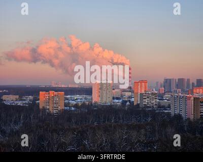Sunset bathes city skyline in warm hues. Smoke billows from industrial chimney into sky. Snow-dusted trees frame urban buildings. Red-and-white striped tower rises amid structures Stock Photo