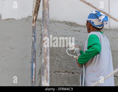 Builder standing on scaffolding while spraying wet concrete plaster on an exterior wall during renovation work. Stock Photo