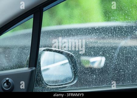 Close-up view from inside a car through a dusty side window with dried spots, showing the exterior mirror and reflecting traffic, illustrating car mai Stock Photo