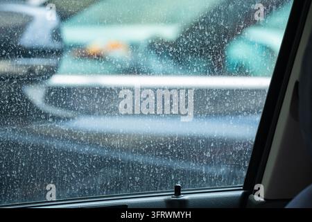 Close-up view from inside a car through a dusty side window with dried spots, showing the exterior mirror and reflecting traffic, illustrating car mai Stock Photo