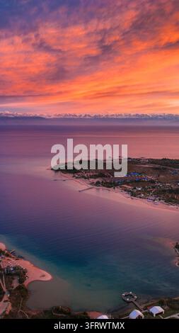 Vertical view of fiery orange and pink sunset clouds reflected across vast calm lake water and distant shoreline Stock Photo