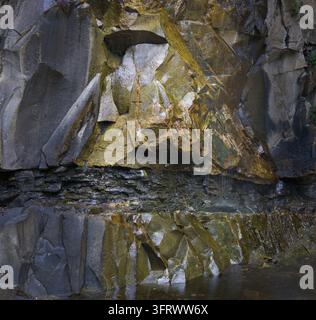Angular blocks of dark wet rock form a natural wall face, marked by yellow green moss and a thin film of water that drips toward a still pool below. Stock Photo