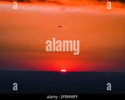 Low sun on the dark horizon beneath orange and red sky with a small dark cloud Stock Photo
