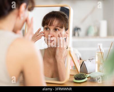 Careful middle-aged lady viewing her complexion in a spacious mirror while seated at a kitchen-table in her house Stock Photo