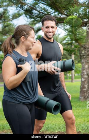 Athletic man and woman in sportswear having a conversation in a green park while holding water bottles and rolled exercise mats. Fitness partners enjo Stock Photo