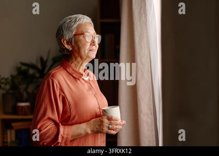 Senior Asian Woman Holding Cup and Looking out Window Stock Photo