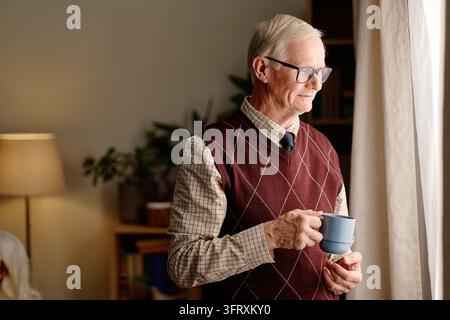 Senior Man Holding Cup Looking through Window at Home Stock Photo