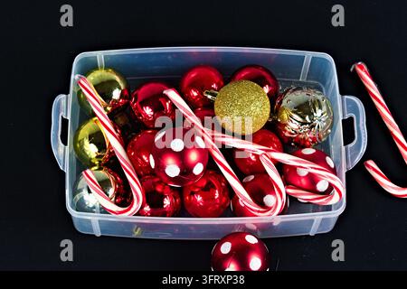 A clear storage bin holds shiny red and gold ornaments, glittery accents, and striped candy canes against a dark background for holiday decorating. Stock Photo