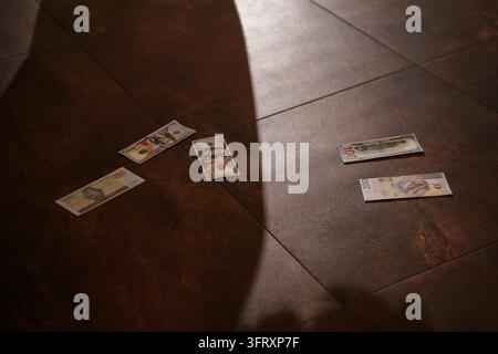 Small play money scattered across a dark brown tiled floor, cast in dramatic sharp shadows, evoking themes of financial risk, gambling, or games Stock Photo