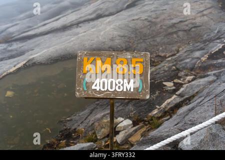 Rugged stone path beside weathered wooden sign. Sign reads "KM 8.5" and "4008M". Signpost stands near misty, rocky terrain. Mountains loom faintly beh Stock Photo