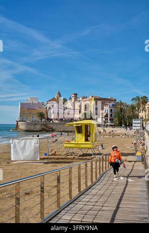 Sitges, Spain - October 10, 2026: A woman walks along the wooden promenade overlooking the sandy beach during a sunny day in the coastal town of Sitge Stock Photo