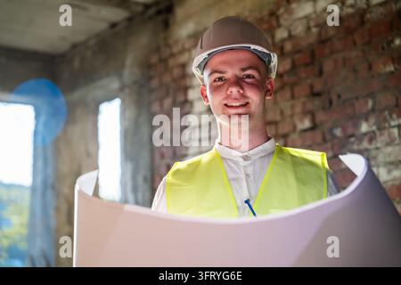 Construction professional in hard hat and high visibility vest reviewing technical drawings inside unfinished building. Architectural planning constru Stock Photo