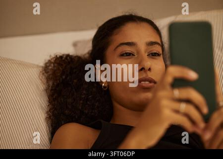 Biracial Woman Scrolling on Smartphone While Lying in Bed Stock Photo