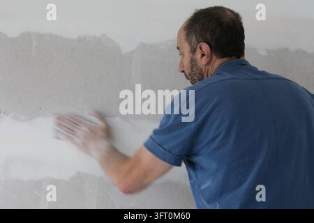 Senior man smoothing dried joint compound on drywall with sandpaper to prepare the wall for painting. Stock Photo