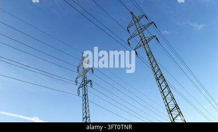 Low-angle view of two steel lattice electricity pylons carrying high-voltage power lines against a clear, deep blue sky with faint wispy clouds and Stock Photo