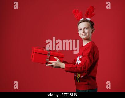 Boy dressed red sweater and reindeer antlers holding wrapped gift box smiling cheerfully happy holiday celebration fun. Stock Photo