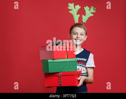 Happy child holding three Christmas gift boxes wearing reindeer antlers and festive sweater smiling bright red background holiday spirit. Stock Photo