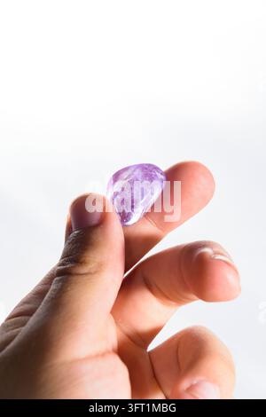 A hand holding a translucent purple amethyst tumbled stone with natural inclusions, lit by bright natural light. Stock Photo