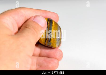 A hand holding a golden brown tiger's eye tumbled stone with dark banding, photographed in natural light against a white background. Stock Photo