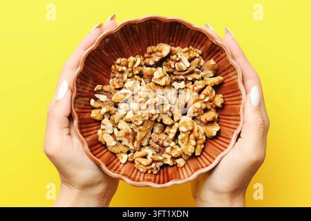 Hands holding brown bowl filled with walnut halves on bright yellow background. Overhead food composition for healthy snacks and natural nutrition. Stock Photo