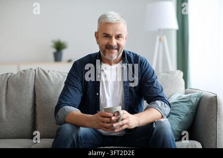 Cheerful senior man with mug of coffee sitting on couch in living room, relaxing at home, copy space. Happy matured bearded grey-haired man resting al Stock Photo