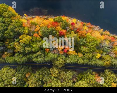 Aerial view of Mount Wachusett surrounded by vibrant autumn foliage with a road winding through the colorful forest near a dark lake in Princeton, Mas Stock Photo