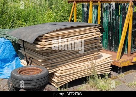 Large stack of flat wooden table tops covered with black tarpaulin, stored alongside old tyres and blue plastic sheeting in an outdoor farm yard setti Stock Photo