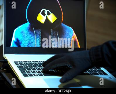 A conceptual technology photograph showing a person in gloves operating a white laptop keyboard, with a screen displaying a hooded figure silhouette a Stock Photo