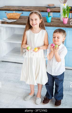 Happy kids holding bright Easter eggs while standing near rustic kitchen counter with flowers and baskets. Stock Photo