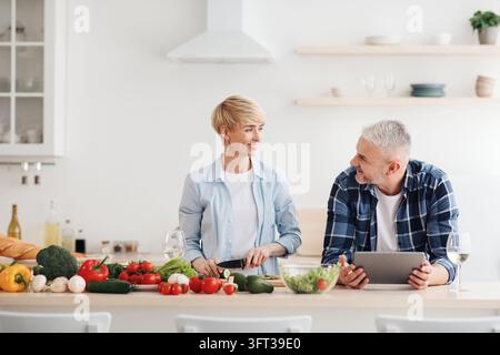 Happy asian family couple cooking food, vegetable salad in kitchen together at home with device. Man and woman culinary according online class to reci Stock Photo