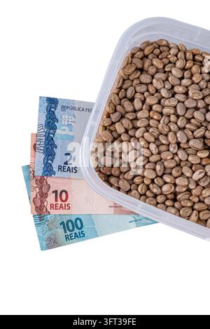 A clear plastic container filled with pinto beans is placed next to several Brazilian Real banknotes, including denominations of 2, 10, and 100. Stock Photo